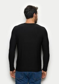 Czarna koszula z długim rękawem, uszyta z gładkiego materiału, z okrągłym dekoltem i dopasowanym krojem, noszona z niebieskimi dżinsami.