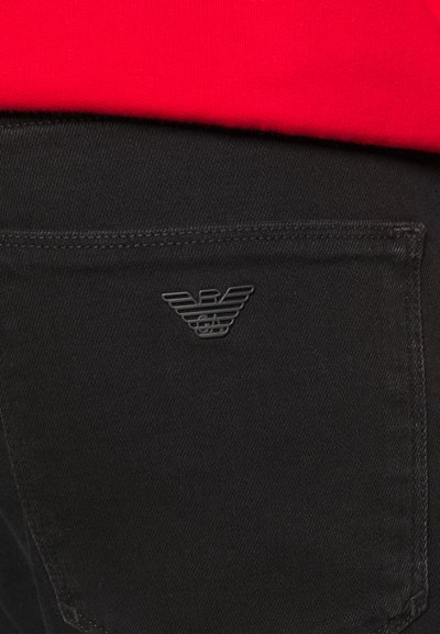 Crne traperice od denima s stražnjim džepom s teksturiranim, reljefnim logotipom. Pojas je djelomično vidljiv, prekriven crvenom majicom.