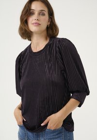 Blusa de terciopelo morado con rayas verticales, mangas cortas abullonadas y un escote acanalado. Llevada con las manos en los bolsillos; la parte superior tiene un corte holgado.