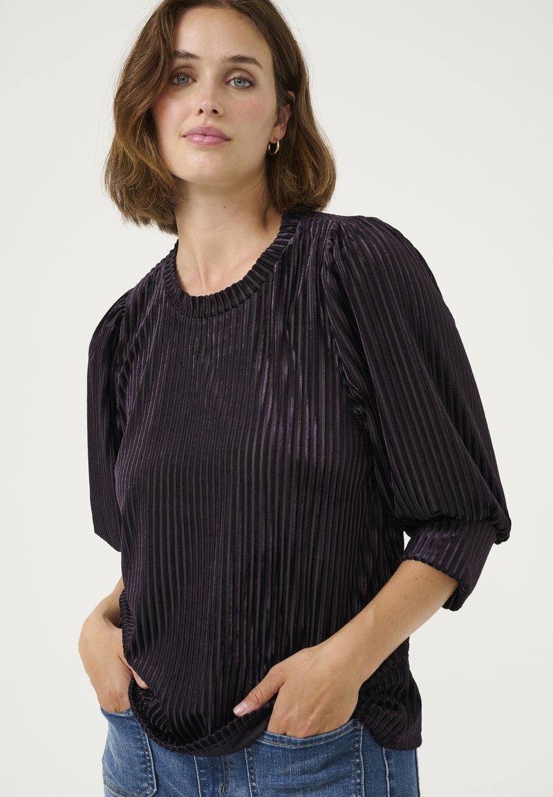 Blusa de terciopelo morado con rayas verticales, mangas cortas abullonadas y un escote acanalado. Llevada con las manos en los bolsillos; la parte superior tiene un corte holgado.