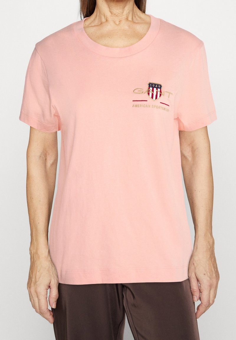 T-shirt en coton rose clair avec un col rond, des manches courtes et un logo brodé sur la poitrine. Texture lisse avec une coupe ample.