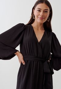 Robe noire en tissu texturé, dotée de longues manches bouffantes, d'un décolleté en V profond et d'une ceinture à nouer à la taille. Design simple et élégant.