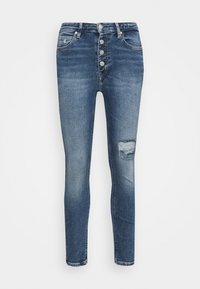 Jeans ajustados de color azul oscuro con cierre de botones, diseño de cinco bolsillos, ligero desgaste y un parche desgastado en el muslo derecho.