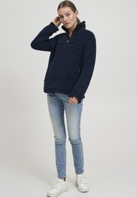 Pullover in pile blu navy con colletto zip nero, superficie testurizzata, tasca frontale e maniche lunghe, abbinato a jeans skinny blu chiaro.