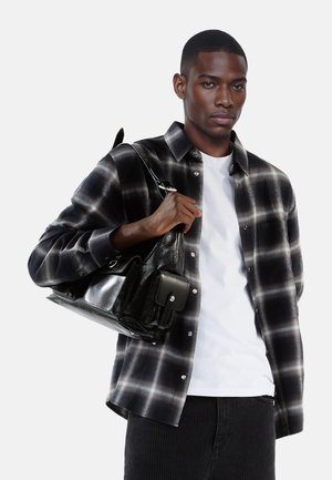 Chemise à carreaux noire et grise avec des boutons, t-shirt blanc en dessous, associée à un sac en cuir noir structuré avec plusieurs poches.