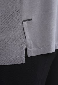 Camiseta gris con un patrón de malla texturizada en el lado izquierdo y tela suave en el derecho. Costuras negras acentúan el dobladillo y la costura lateral.