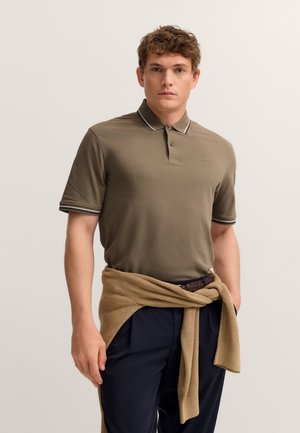 Olivgrünes Poloshirt mit Kragen und einem hell gestreiften Besatz. Kombiniert mit marineblauen Hosen und einem beigen Pullover, der in der Taille gebunden ist.