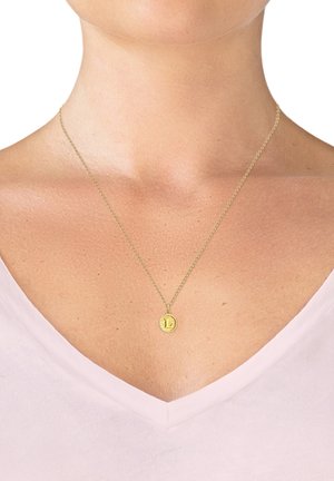 Goldene Anhänger-Nackenlace an einer dünnen Kette, mit einem texturierten runden Charm und einem eingravierten "L." Getragen über einem hellrosa V-Ausschnitt-Oberteil.