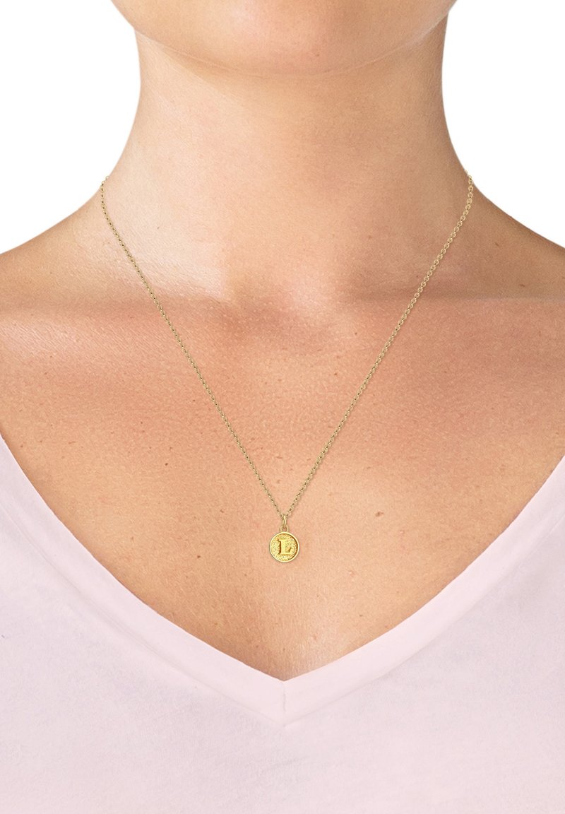 Guldvedhæng på en tynd kæde, prydet med en tekstureret rund charme med et indgraveret "L." Båret over en lys pink V-hals top.