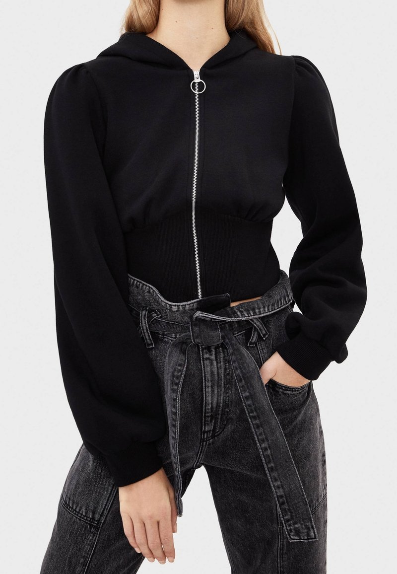 Felpa corta nera con zip frontale, dettagli in argento e maniche a sbuffo, abbinata a pantaloni in denim scuro a vita alta con cintura annodata.