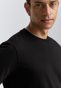 Sort kortærmet t-shirt lavet af blødt, strækbart stof; rund halsudskæring og glat overflade. Nærbillede af bryst- og skulderområde.