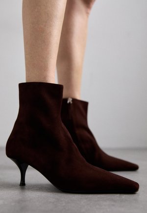 Botas de ante marrón hasta el tobillo con punta afilada, tacón alto elegante y detalle de cremallera en la parte posterior, con una textura suave y un diseño minimalista.