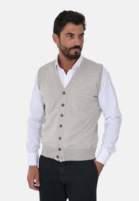 Gilet lavorato a maglia grigio chiaro con scollatura a V, dotato di sei bottoni scuri e un design attillato, indossato sopra una camicia bianca a maniche lunghe.