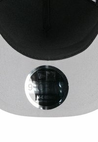Schwarze und graue verstellbare Kappe mit glatter Oberfläche, ausgestattet mit einem runden metallischen Etikett, das "9FORTY M-CROWN" beschriftet ist. Die Innenseite zeigt keine zusätzlichen Muster.