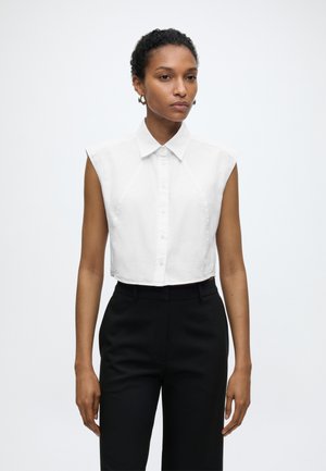 Mujer con una camisa corta blanca sin mangas con botones y pantalones negros de talle alto, de pie frente a un fondo liso y claro.