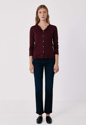 Donna in piedi con cardigan bordeaux abbottonato, jeans blu scuro a gamba dritta e mocassini neri su uno sfondo chiaro e uniforme.