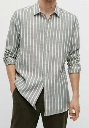 Homme portant une chemise boutonnée à rayures verticales gris et blanc, manches retroussées, associée à un pantalon foncé, main dans la poche.