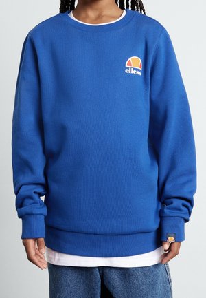 Person iført en knaldblå Ellesse-sweatshirt over en hvid skjorte og blå jeans, med logo synlig på brystet og håndleddet.