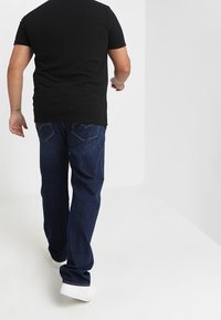 Musta lyhythihainen paita, siniset farkut takataskuilla, valkoiset tennarit, nähtynä takaa vaaleaa taustaa vasten.