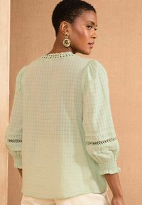 Blusa verde chiaro con motivo a griglia, maniche a sbuffo e bordo in pizzo sui polsini. Vista posteriore visibile su uno sfondo neutro.