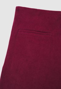 Primo piano di tessuto in corduroy bordeaux con una texture a righe verticali e un dettaglio di tasca a filetto orizzontale.