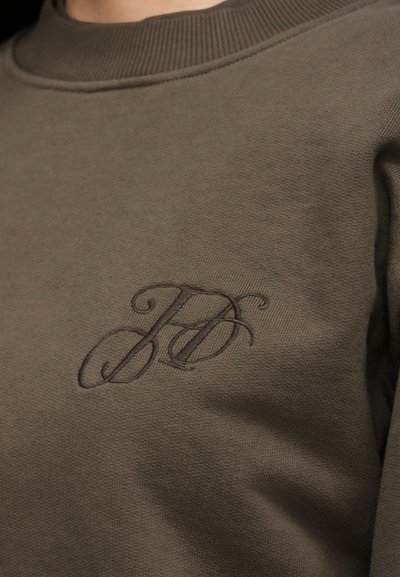 Κοντινό σε καφέ φούτερ με λεπτεπίλεπτο κεντημένο λογότυπο με επικαλυπτόμενα αρχικά "JD" στο στήθος και ραβδωτό λαιμόκοψη.