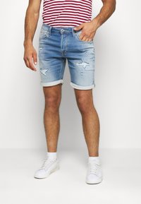 Mężczyzna w jasnoniebieskich, zniszczonych szortach dżinsowych z podwiniętymi nogawkami, w czerwono-białą w paski koszulę i białe trampki, stojący na białym tle.