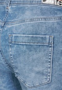 Light blue denim jeans feature a back pocket with contrasting stitching and a textured finish. Logo tag visible at the waistband.