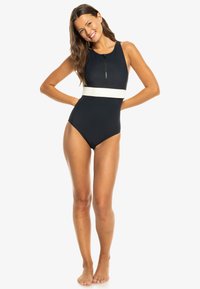 Laivastonsininen yksiosainen uimapuku, jossa on valkoinen vaakasuora korostusnauha. Varustettu etuvetoketjulla ja ilman hihoja, siinä on sileä materiaali.