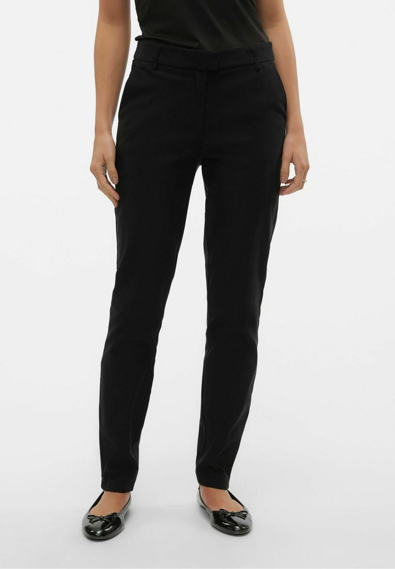 Femme portant un pantalon noir ajusté, une chemise noire et des ballerines noires, debout devant un fond clair uni.