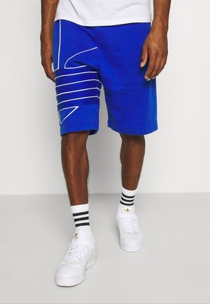 Blauwe sportshorts van katoen, met witte lijnaccenten en geometrische ontwerpen aan de zijkant; gedragen met witte sneakers en gestreepte sokken.