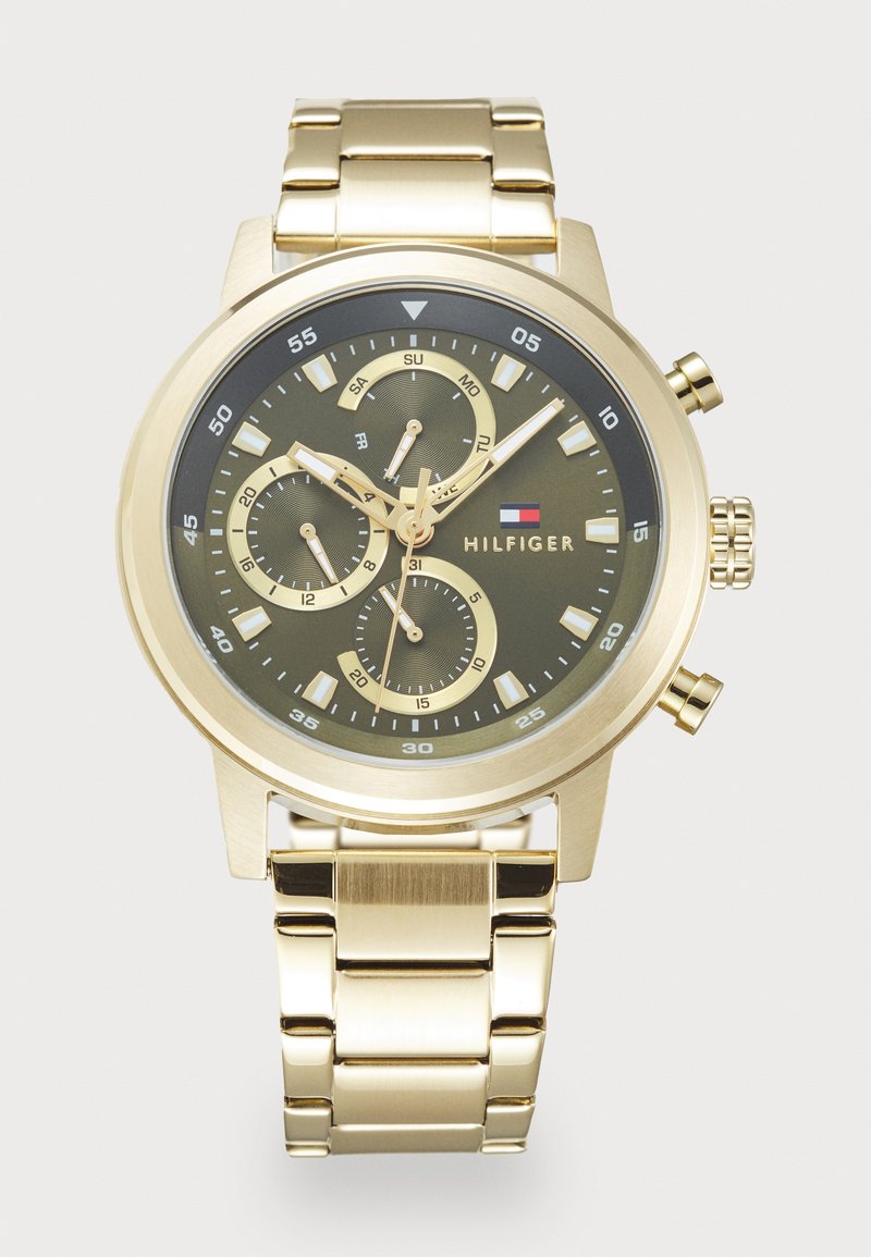 Montre-bracelet en métal doré avec un cadran sombre, trois sous-cadrans indiquant les jours, l'heure sur 24 heures et les secondes, ainsi que le logo Tommy Hilfiger sur le cadran.