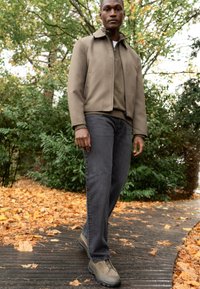 Man som går på en lövbelagd träpromenad i grön jacka, mörka jeans och beige skor, omgiven av höstträd och buskar.