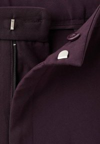 Gros plan sur un pantalon violet foncé avec une fermeture éclair, un bouton et un système de crochet et de barre en métal à la taille.