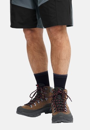 Bruine wandelschoenen met zwarte accenten en rode veters, gedragen met zwarte enkelsokken. De korte broek is donkergrijs met een gelaagd ontwerp.