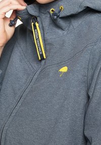 Grå zip-jacka med hög krage, gula accenter och en liten paraplylogotyp. Har justerbara snoddar och texturerad tyg.