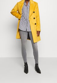 Gul ullblandad kappa med notchske collar, fyra svarta knappar, bärs över en svart- och vitrandig skjorta och grå smala jeans med ankelboots.