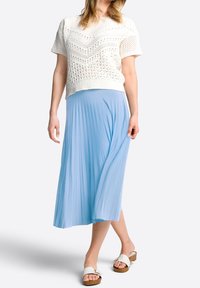 Donna che indossa un top a maglia bianco a maniche corte, una gonna midi plissettata azzurro chiaro e sandali bianchi aperti davanti con tacco in legno, su sfondo bianco.