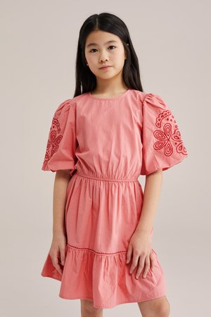 Joven con cabello largo y negro, vestida con un vestido rosa de mangas abullonadas y bordado floral rojo en las mangas.