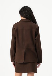 Hnedý semišový blazer so štruktúrovaným dizajnom, dlhými rukávmi a zadným rozparkom, doplnený o zodpovedajúcu hnedú semišovú sukňu.