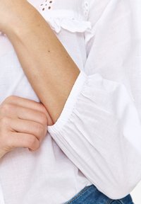 Biała lniana bluzka z bufkami i marszczonymi mankietami, z teksturowanym wykończeniem. Kolor skóry widoczny w pobliżu mankietu bluzki.