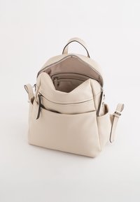 Sac à dos en cuir beige avec un compartiment principal zippé, une poche avant zippée et des bretelles réglables, mettant en valeur une texture lisse.