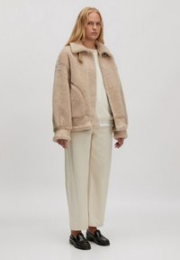 Beige schapenvachtjack met een kraag, zijzakken en een rits. Ggedragen over een crèmekleurige trui en gecombineerd met lichte, rechte broeken. Zwarte instappers.