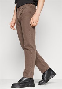 Mies, jolla on päällään ruskeat suoralahkeiset farkut ja mustat nahkaiset nyörisaappaat, seisoo jalka hieman kohotettuna yksivärisellä taustalla.