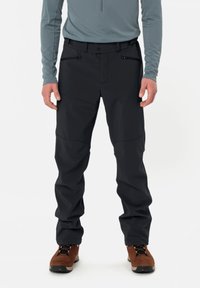 Svarta utomhusbyxor med skräddarsydd passform, med dragkedjor på fickorna och förstärkta knän. Kombineras med en grå långärmad tröja och bruna stövlar.