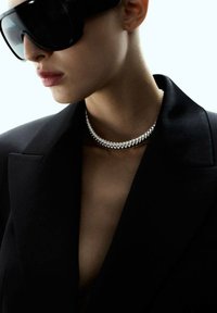 Femme portant de grandes lunettes de soleil noires, un collier en argent massif et un blazer noir avec un décolleté ouvert.
