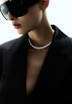 Donna che indossa grandi occhiali da sole neri, una collana d'argento massiccia e un blazer nero con colletto aperto.