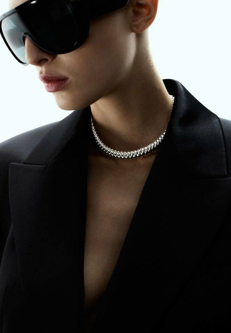 Femme portant de grandes lunettes de soleil noires, un collier en argent massif et un blazer noir avec un décolleté ouvert.