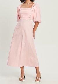 Vaaleanpunainen midi-mekko puffipolloilla, neliönmuotoisella pääntiellä ja vaakasuorilla poimuilla vartalossa. Kankaan sileä tekstuuri, yksinkertainen muotoilu.