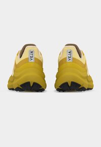 Ζευγάρι κίτρινων αθλητικών παπουτσιών Veja, εμφανιζόμενα από πίσω, με παχιά σόλα με μαύρη υφή και ανακλαστικές ετικέτες στη φτέρνα με το όνομα της μάρκας.